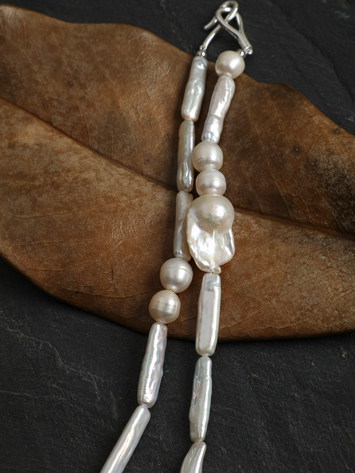 A beaded necklace with irregular white baroque pearls and smooth white beads, featuring a large central pearl and a silver clasp, displayed on a brown dried leaf over a dark slate background.
